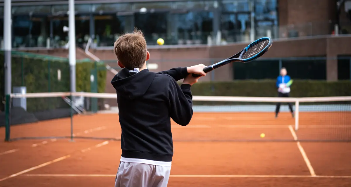A junior boy hits a forehand with the Saber during a rally with his coach on a clay court